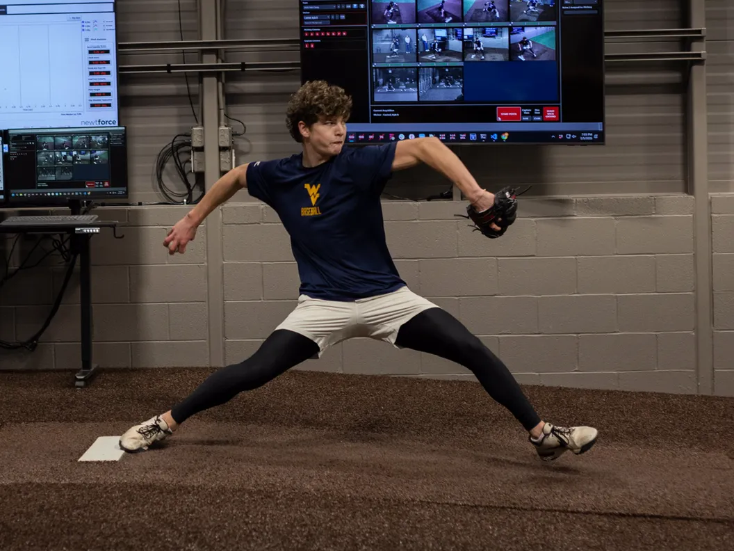 A WVU Baseball pitcher mid-delivery on the mound in the biomechanics lab, with motion-capture and force-plate screens on the wall behind him